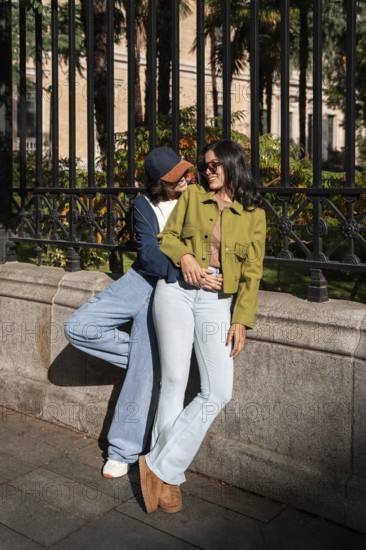 A joyful lesbian couple stands close, smiling and holding hands in a sunlit urban setting. They radiate happiness and togetherness, enjoying each other's company