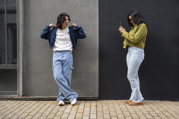 A lesbian couple captures a candid moment outdoors. Both appear happy and relaxed in casual attire against a two-tone wall, sharing a joyful connection