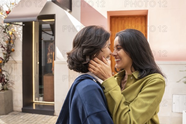 A lesbian couple smiling warmly as they embrace outdoors. The atmosphere is sunlit and joyful, reflecting love and connection. Background features a modern, stylish facade