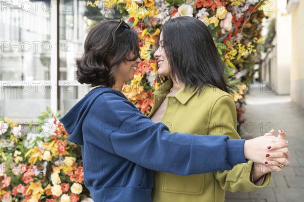 A lesbian couple shares an affectionate moment, embracing amidst a colorful flowered wall. Their expressions radiate joy and connection, symbolizing love and unity