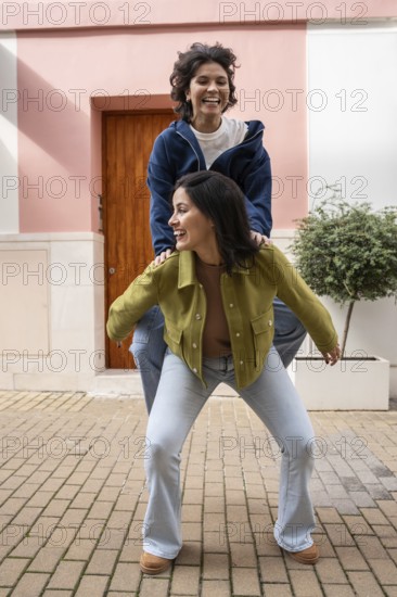 A joyful lesbian couple shares a playful moment outdoors, with one partner giving the other a piggyback on a city street. They smile, enjoying a carefree day together