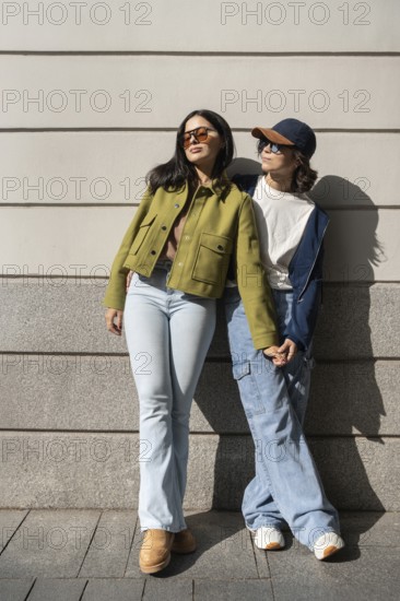 A lesbian couple stands outside, holding hands and enjoying the sunlight. Dressed in casual attire, they exude a sense of happiness and relaxation against a wall in an urban setting