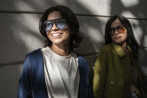 A lesbian couple enjoying the outdoors, wearing stylish sunglasses and jackets. They stand against a white wall under patches of light, smiling warmly and exuding a joyful vibe