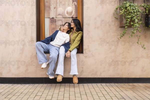A lesbian couple sits affectionately on a windowsill, holding hands and enjoying a heartfelt moment. The background features a blush-toned wall and a hanging plant