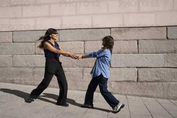 A joyful multiethnic lesbian couple holds hands and dances outside against a stone wall, celebrating love and pride in a lively urban setting