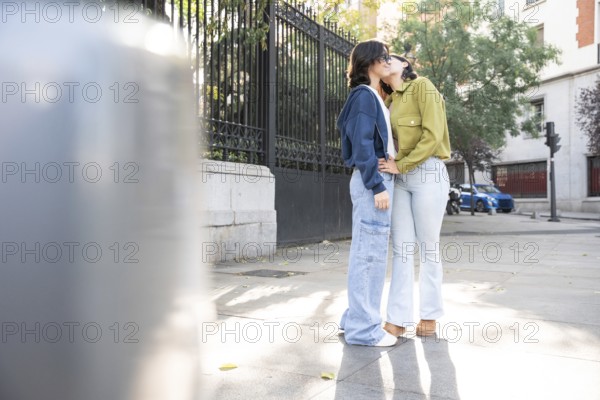 A loving lesbian couple enjoying a sunny day on a city street. They are embracing and sharing a kiss, surrounded by urban architecture and greenery