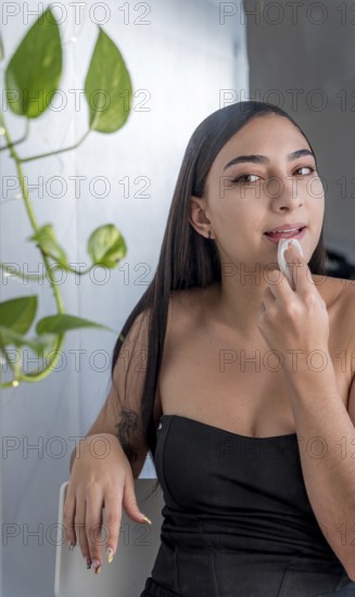 A woman is seated, applying makeup with a sponge in a softly lit room. She wearing a black top, with a green plant visible in the background, creating a serene atmosphere