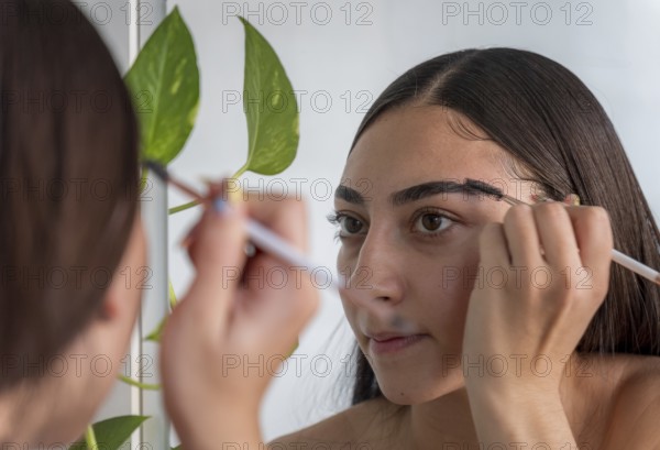 A woman applies makeup with a brush, concentrating on her reflection in a mirror. A green plant adds a natural touch to the setting, emphasizing beauty and care