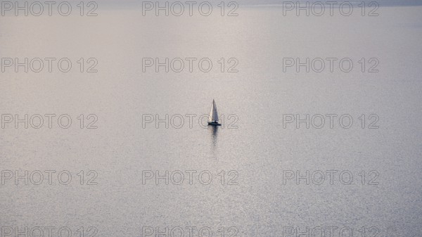 A solitary sailboat on the tranquil waters of Lake Geneva during a sunny winter day, with the snowy Alps looming in the distance, captured from the scenic Lavaux vineyards