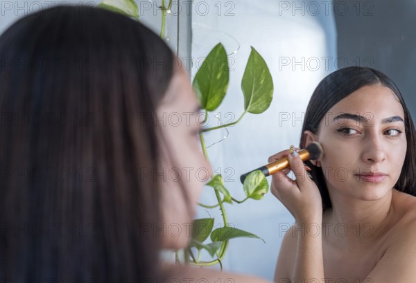 A woman is applying makeup with a brush while looking into a mirror. Green plants add a natural touch to the serene setting, creating a calming atmosphere