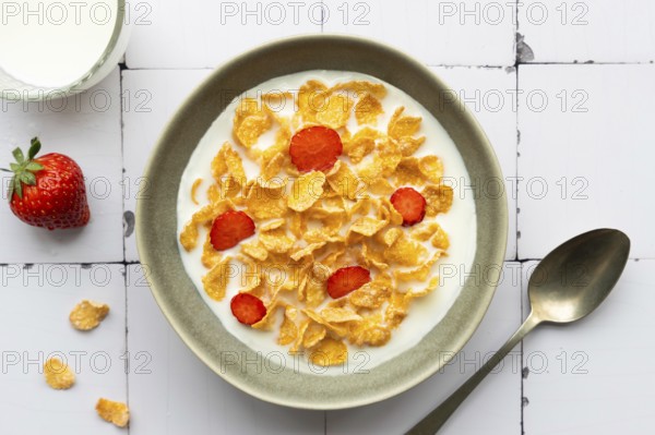 A bowl of crunchy cornflakes with fresh strawberry slices, served in milk. A wholesome breakfast setting with a glass of milk and a spoon placed nearby