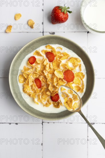 Top down view of a bowl with cornflakes and strawberry slices in milk, with a spoon resting inside. A fresh strawberry and a glass of milk sit nearby on the surface