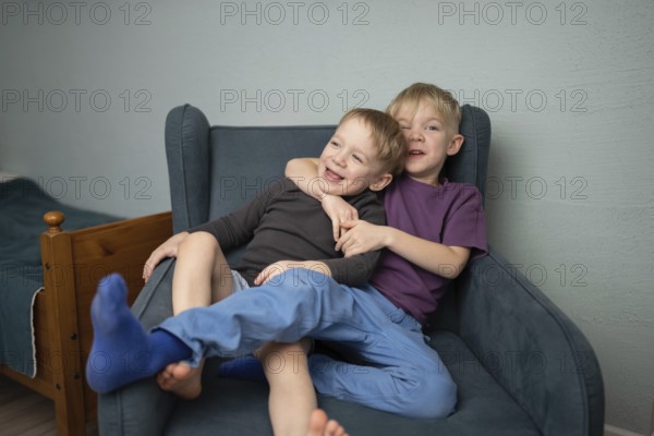 Two joyful young brothers share a warm embrace on a cozy blue armchair, radiating happiness and sibling love in their bedroom. A perfect depiction of brotherly affection