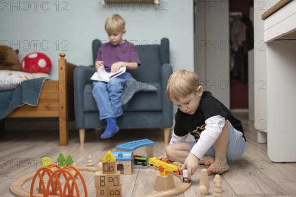 Two young children in a cozy room, one plays with wooden train toys on the floor, while the other sits in a chair reading a book, surrounded by toys and playful decor