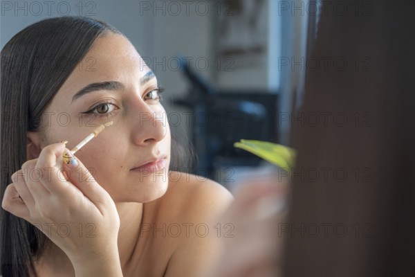 A woman applies concealer under her eyes using a makeup wand, focusing on her reflection in the mirror. The setting is relaxed, emphasizing natural beauty and care