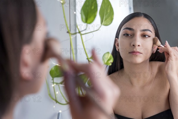 A woman carefully applies makeup using a brush in front of a mirror for the makeup session. Her reflection captures her focused expression, with green plant decor enhancing the serene ambiance
