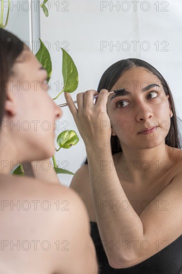 A woman focuses on applying makeup with a brush in front of a mirror for the makeup session. A green potted plant decorates the reflective background, adding a natural touch to the space