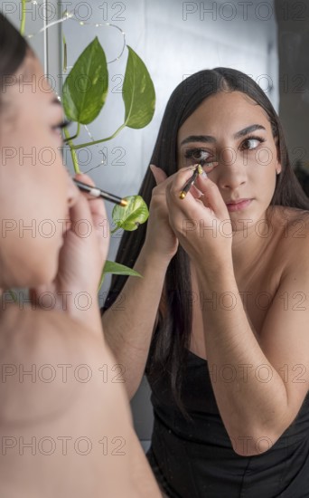 A woman in a black outfit applies makeup in front of a mirror. She holds a makeup brush near her eye, focusing intently, with green plant leaves nearby