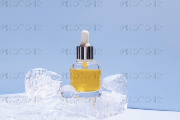 A serum bottle with a dropper, filled with yellow fluid, delicately placed amongst transparent ice chunks against a uniform blue backdrop, highlighting the freshness or cooling aspect of the product