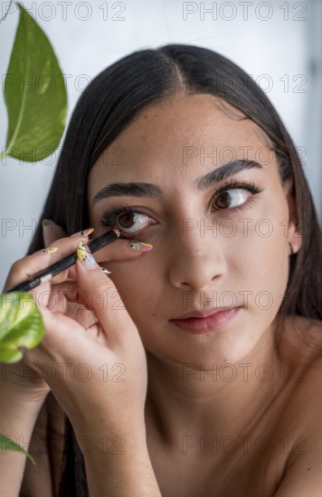 A woman applies eyeliner with precision, focusing intently. Her delicate hand grips the pencil, showcasing neat manicured nails. Natural light illuminates her serene expression