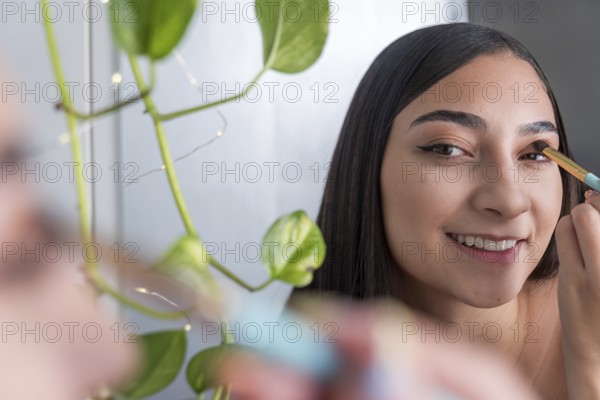 A woman is seen smiling while applying eye makeup in front of a mirror for the makeup session. A green plant is visible, adding a touch of nature to the scene and enhancing the composition