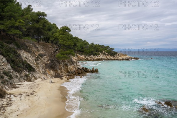 Mega Portokali Beach in southern Greece features a secluded sandy cove with turquoise waters and rocky cliffs covered in pine trees. A tranquil coastal landscape under a cloudy sky