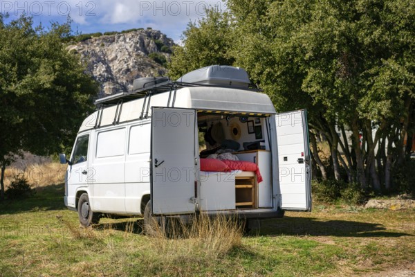 A cozy camper van with open doors reveals a comfortable interior, set against a backdrop of lush greenery and rocky hills in Greece, ideal for adventure seekers