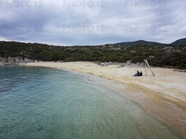 A peaceful aerial view of Kalamitsi wild beach in Greece. A couple sits on the golden sand by turquoise waters, surrounded by lush greenery and a serene natural landscape under a cloudy sky