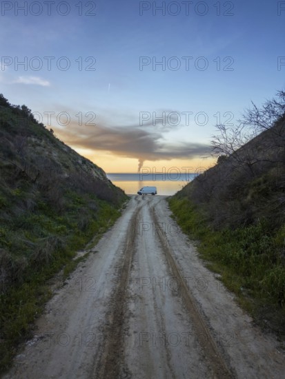 A lone camper van is parked on a dirt path leading to a tranquil Greek seaside. The sky is painted with soft sunset hues, creating a peaceful, scenic atmosphere