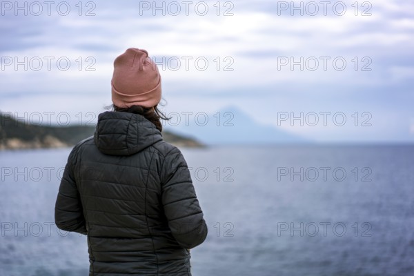 A person in a pink beanie and black jacket stands by the sea in Greece, gazing at the horizon. The serene ocean view is framed by distant mountains under a cloudy sky