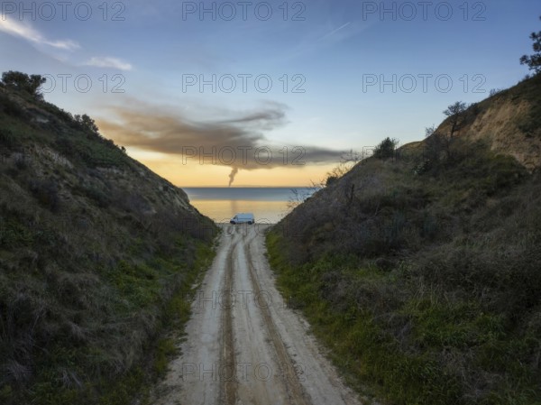 A narrow road flanked by green hills leads to a tranquil beach in Greece. A van is parked near the shore, with a calm sea and vibrant sunset painting the horizon
