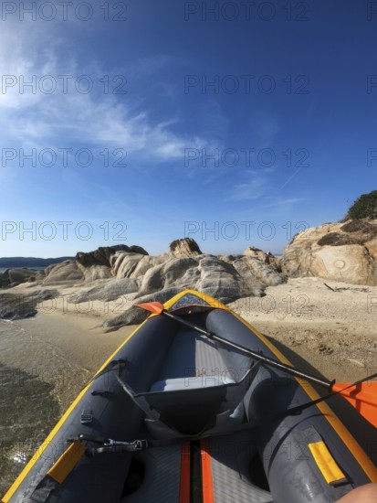 Inflatable kayak resting on a sandy shore in Greece. The vibrant kayak contrasts beautifully with the clear blue sky and rugged coastal rocks. Perfect for outdoor adventure themes