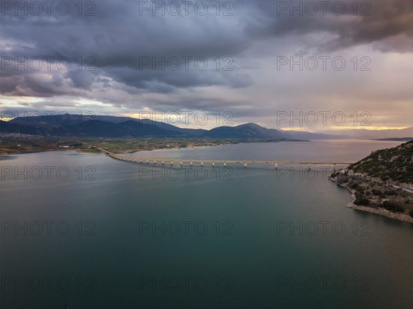 A stunning aerial view of Lake Polyfyto with the Servia Neraida high bridge spanning the water. Surrounded by mountainous landscapes, the scene captures the beauty of sunset and dramatic skies