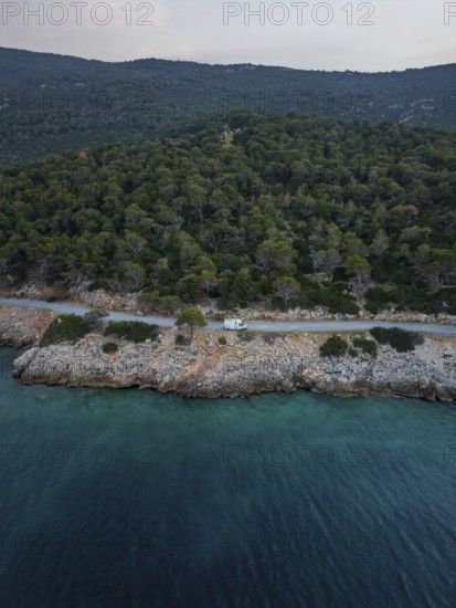 Aerial view of a van in a coastal road running through lush green forest by a turquoise sea in Greece. The serene landscape shows harmony between nature and adventure