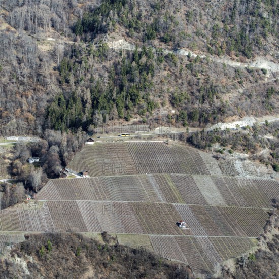 A breathtaking aerial view of Swiss farmland with neatly arranged fields against a rugged hillside. The earthy tones and sparse vegetation suggest a late autumn or early winter season