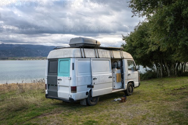 Camper van parked beside a scenic lake in Greece, surrounded by lush greenery and mountains. Cloudy sky sets a tranquil mood for travel and adventure in nature, offering relaxation
