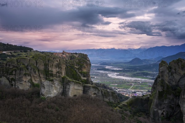Captivating sunset view of the historic Holy Trinity Monastery perched atop towering cliffs in Greece, with a serene valley and distant snow-capped peaks under a dramatic sky