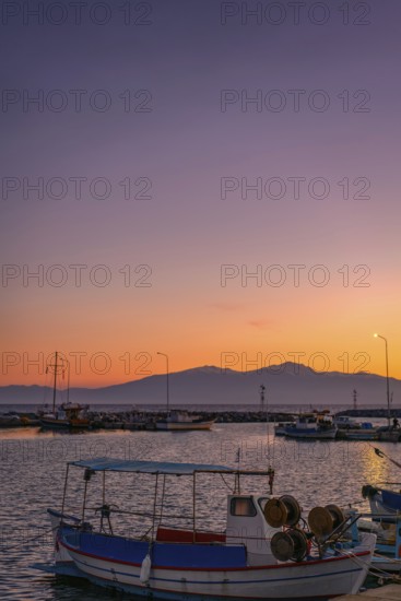 Angelochori port in Greece at sunset, featuring colorful fishing boats in the marina. Mount Olympus stands majestically in the background, framed by warm orange and purple skies