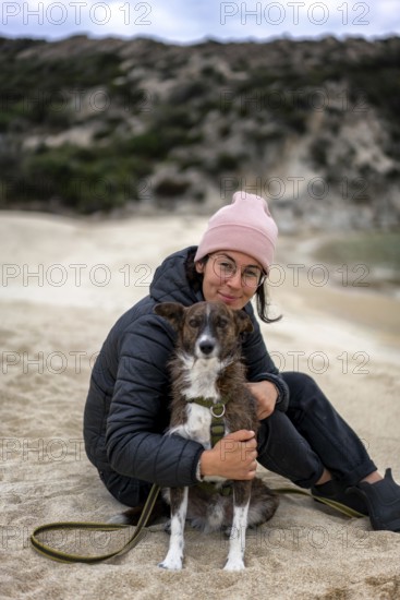 A woman wearing a pink beanie and glasses sits on a sandy beach in Greece, hugging a dog. The relaxed atmosphere captures a cozy moment in nature