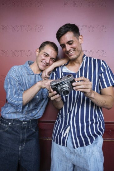 Charming gay couple smiling while reviewing photos on a vintage camera against a red wall. Casual outfits, relaxed poses, and warm interaction reflect joy and a shared passion for photography