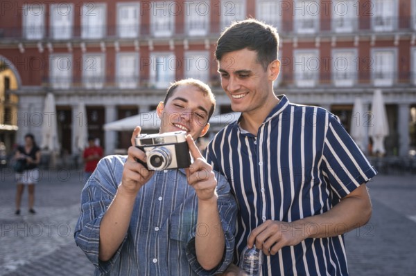 Happy gay couple reviewing photos on a vintage camera in Madrid's Plaza Mayor. Casual fashion, sunny weather, and historic architecture create a joyful and relaxed travel moment