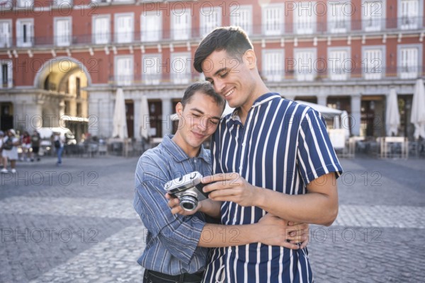 Gay couple embracing while reviewing photos on a vintage camera in Madrid's Plaza Mayor. Warm smiles, casual fashion, and historic architecture capture a romantic and joyful travel moment