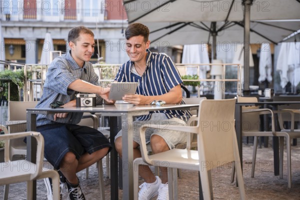Gay couple sitting at an outdoor cafe in Plaza Mayor, Madrid, smiling while using a tablet and sharing photos from a vintage camera. Relaxed atmosphere with casual fashion and city vibes