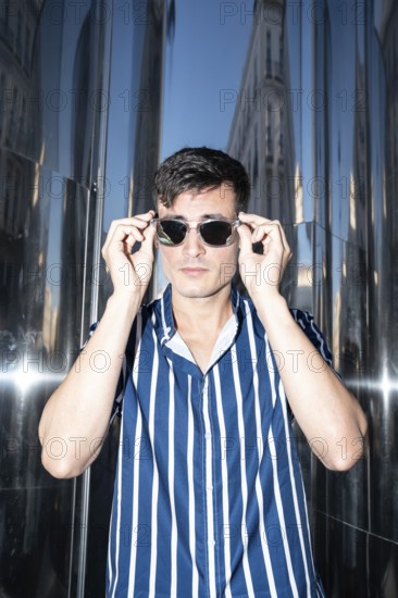Fashionable young man in a striped shirt adjusting his sunglasses while posing against a reflective metal background in the city. Confident, urban, and stylish vibe