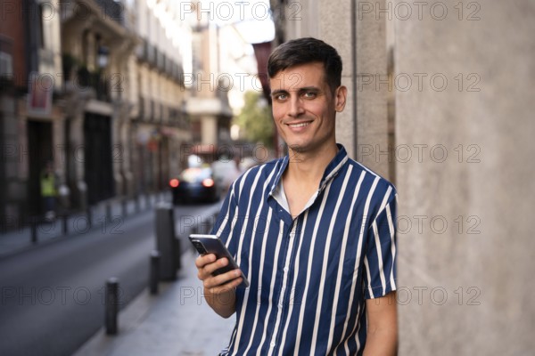 Young man in a striped shirt holding a smartphone and smiling on a city street. Relaxed and stylish, he stands against an urban backdrop, enjoying a modern and casual moment