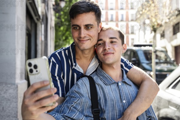 A joyful gay couple takes a selfie while embracing on the lively streets of Madrid. Their expressions reflect love and pride, celebrating togetherness in an urban setting