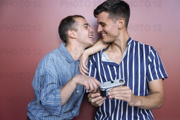 Joyful gay couple sharing a candid moment while holding a vintage camera against a colorful wall. Casual fashion, genuine smiles, and playful interaction highlight their bond and happiness