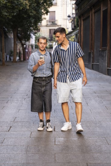 Stylish gay couple walking hand in hand on a quiet street in Madrid, Spain. One checks their smartphone while they enjoy a relaxed and casual stroll in the warm evening light