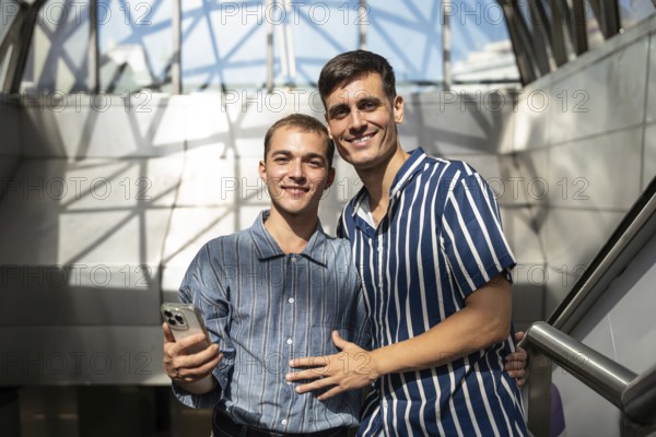 Gay couple smiling and enjoying a sunny day on the streets of Madrid, celebrating pride and love. Urban setting with modern architecture in the background