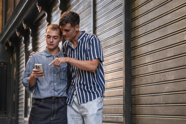 Stylish young gay couple standing together on a city street in Madrid, Spain, sharing a moment while using a smartphone. Casual fashion and urban surroundings create a warm vibe